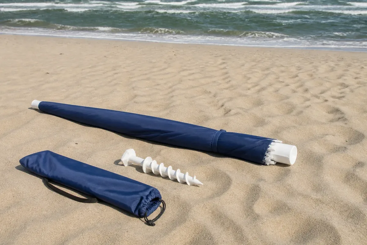 Closed beach umbrella lying safely on sand beside a screw-in anchor and carry bag